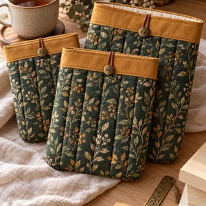 May include: Two rectangular quilted fabric sleeves featuring a dark green background with a botanical print of leaves and small flowers. Each sleeve has a solid mustard yellow top band, a vertical quilted pattern, and a single button closure.