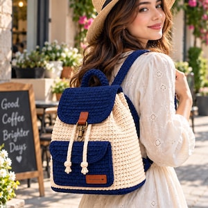 May include: A crochet backpack featuring a two-tone design with a dark navy blue top flap, base, and front pocket, contrasted by a cream-coloured main body. It includes a brown leather-like tab, a brass-toned metal clasp, and cream drawstring cords.