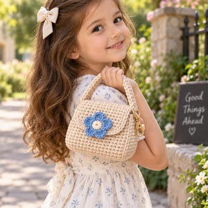 May include: A beige crocheted handbag featuring a single blue flower applique with a white center on the front flap. The bag has a sturdy crocheted handle attached with gold-toned metal swivel clasps, providing a structured and textured appearance.