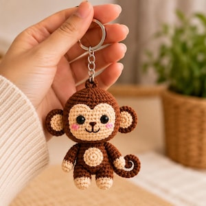 May include: A brown and beige crocheted monkey keychain featuring a round head with black eyes, pink cheeks, a small embroidered smile, and a curled tail. The monkey is attached to a silver-colored metal split ring by a short chain link.