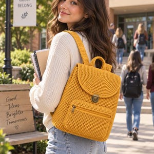 Puede incluir: Una mochila de ganchillo color amarillo mostaza con una solapa estructurada, un cierre de presión de metal color bronce, un asa superior, correas ajustables para los hombros y un bolsillo frontal con cremallera y tirador de metal bronce.