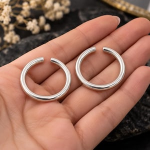 May include: Two circular silver-toned open rings with a smooth, polished finish. Each ring features a thick, rounded band design with a small gap at the top, creating a continuous C-shaped silhouette that is uniform in thickness and metallic luster.