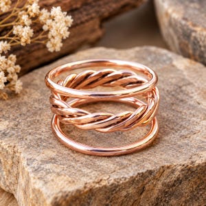 Op de afbeelding: Een stapelbare ringenset bestaande uit drie afzonderlijke ringen in een gepolijste roségouden tint. De set bevat twee gladde, effen ringen en een middelste ring met een gedraaide, touwachtige textuur, allemaal met een reflecterende afwerking.