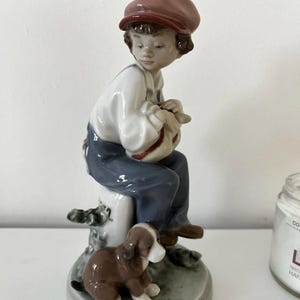 Puede incluir: Una figura de porcelana de un niño con gorra marrón, camisa blanca y peto azul, sentado sobre un poste blanco mientras sostiene un objeto pequeño. Un cachorro marrón y blanco está sentado en la base del poste sobre una peana gris circular.