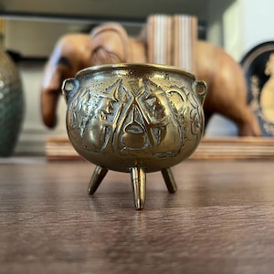 May include: A small, round, three-legged brass cauldron featuring embossed imagery of two figures wearing pointed hats standing on either side of a central pot. The metallic surface has a polished finish with darkened recessed details.