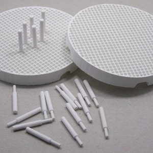 May include: Two white ceramic discs with a grid pattern, one with small white ceramic cylinders on top. A pile of additional white ceramic cylinders are scattered on a gray surface.