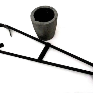May include: A black metal tool with a handle and two loops at the ends. The tool is used to hold a gray ceramic crucible.