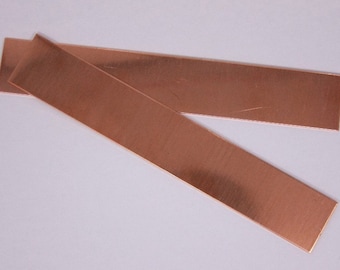 May include: Two thin strips of copper metal, each about 10 inches long and 1 inch wide, are lying on a white surface. The strips are shiny and have a slightly textured surface.