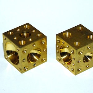 May include: Two brass cubes with multiple drilled holes and rounded interior cutouts. The cubes have a polished, reflective surface and are set against a white background. The cubes are likely used for decorative or artistic purposes.