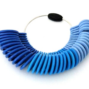 Op de afbeelding: Een set blauwe plastic ringmaten in verschillende blauwtinten, bevestigd aan een zilveren metalen ring met een zwart schuimrubberen deel. De ringmaten zijn in een halve cirkel gerangschikt om vingeromtrekken te meten.