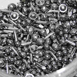 May include: A close-up view of a jar filled with a mixture of small, round, and cylindrical metal shot. The shot is a dark, metallic gray color.