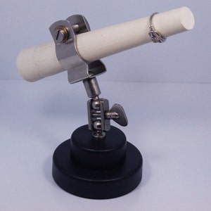 May include: A jewelry display stand with a white cylindrical post held by a metal clamp. The stand has a ball joint and a black circular base. A silver ring with a decorative design is on the post.