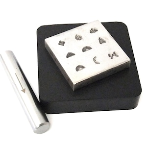 Puede incluir: Un juego de sellos metálicos para la elaboración de joyas, que presenta una variedad de diseños que incluyen corazones, hojas y lazos. Los sellos están dispuestos sobre una base cuadrada negra, y se incluye un martillo de metal.