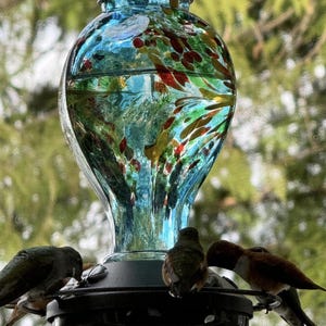 May include: A glass hummingbird feeder with a bulbous, light blue reservoir featuring multicolored flecks of red, yellow, and green. The feeder has a black circular base with multiple feeding ports where several small birds are perched.