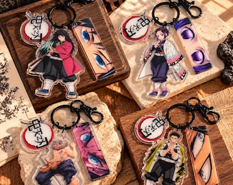 Puede incluir: Cuatro llaveros de acrílico con personajes de anime de Demon Slayer. Cada llavero incluye una figura, una pieza rectangular con un primer plano de ojos y un dije circular con texto en japonés, unidos a anillos de metal negro.