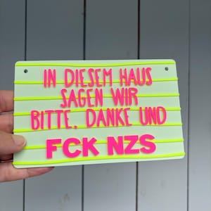 Könnte beinhalten: Ein rechteckiges hellgrünes Schild mit horizontalen gelben Linien und erhabenem neonpinkem Text, der lautet: IN DIESEM HAUS SAGEN WIR BITTE, DANKE UND FCK NZS. Das Schild hat zwei kleine kreisförmige Löcher an den oberen Ecken zur Befestigung.