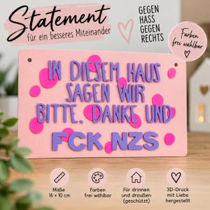 Könnte beinhalten: Ein rechteckiges rosa Schild mit violettem 3D-gedrucktem Text: IN DIESEM HAUS SAGEN WIR BITTE, DANKE UND FCK NZS. Das Schild ist mit verstreuten pinkfarbenen Kreisen verziert und misst laut der Angabe am unteren Rand 16 cm mal 10 cm.
