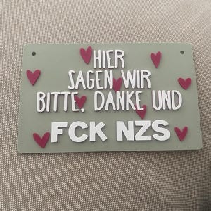 Könnte beinhalten: Ein rechteckiges, salbeigrünes Schild mit weißer, erhabener Schrift, die HIER SAGEN WIR BITTE, DANKE UND FCK NZS lautet. Das Schild ist mit mehreren kleinen, dunkelrosa herzförmigen Akzenten verziert, die um den Text verteilt sind, und hat zwei Löcher.