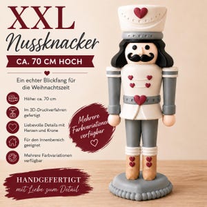 Könnte beinhalten: Eine 70 cm hohe Nussknacker-Figur in cremefarbener Uniform mit dunkelroten Herz-Applikationen auf Hut, Jacke und Stiefeln. Die Figur hat schwarze Haare, einen schwarzen Schnurrbart und steht auf einem runden, grauen Sockel mit Perlenrand.