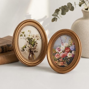 May include: Two oval-shaped gold picture frames with beaded edges. The left frame contains a print of white flowers in a vase, and the right frame contains a print of a colourful bouquet of roses and other flowers in various shades.