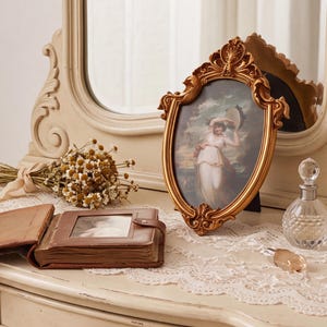 May include: An oval gold-toned ornate picture frame containing a print of a person holding a tambourine, placed on a white lace runner next to a brown leather-bound photo album, a small bouquet of dried chamomile flowers, and a glass perfume bottle.
