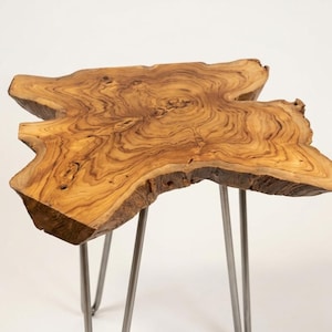 May include: A side table featuring a natural live edge wood slab top with visible brown grain patterns and organic contours. The tabletop is supported by three slender, silver-colored metal hairpin legs that extend downward from the underside.