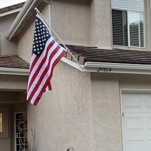 May include: A United States flag with red, white, and blue colors hanging from a white pole mounted to the exterior wall of a beige house. The house features a white garage door, a window with dark shutters, and the house number 29864 displayed.