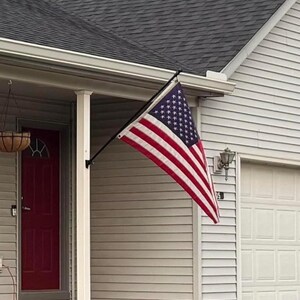 May include: A rectangular American flag with thirteen horizontal red and white stripes and a blue canton featuring fifty white stars. The flag is mounted on a black metal pole attached to a white porch column on the exterior of a house.