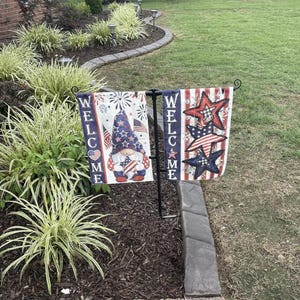 May include: A double-sided garden flag set on a black metal stand. The left flag features a gnome in a star-patterned hat holding a star, with the text WELCOME. The right flag displays three stacked stars in red, white, and blue with the text WELCOME.