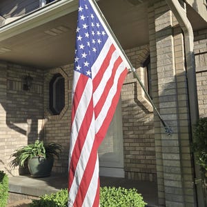 May include: A United States flag featuring a blue canton with white stars and thirteen alternating red and white horizontal stripes, mounted on a silver metal pole attached to a brick exterior wall near a front entrance.