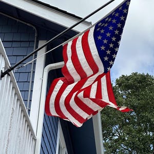 May include: The flag of the United States of America, featuring thirteen horizontal red and white stripes and a blue canton with white stars, is mounted on a black pole attached to the exterior wall of a blue-sided building with white trim.