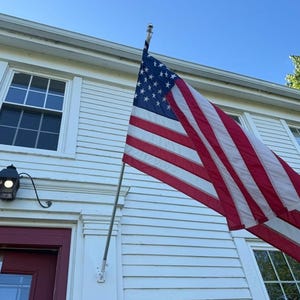 May include: The American flag with red and white stripes and a blue canton featuring white stars is mounted on a silver pole attached to the white horizontal siding of a building exterior with a red door and a black wall-mounted light fixture.