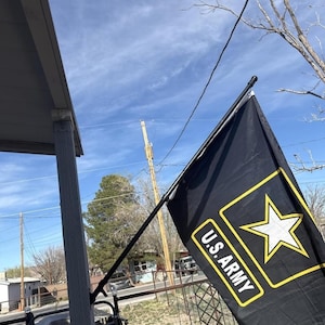 May include: A black rectangular flag featuring a yellow border, a white five-pointed star, and the text U.S. ARMY in white capital letters. The flag is mounted on a black pole attached to a wooden post, displayed against a bright blue sky.