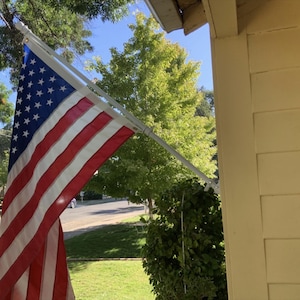May include: A rectangular American flag featuring thirteen horizontal red and white stripes and a blue canton with fifty white stars. The flag is attached to a white metal pole mounted at an angle to the side of a light yellow building exterior.