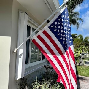 May include: A rectangular United States flag featuring thirteen horizontal red and white stripes and a blue canton with fifty white stars. The flag is attached to a white metal pole mounted to the exterior wall of a building near a window.