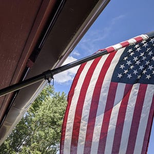 May include: A United States flag attached to a black metal pole mounted on a building exterior. The flag features a blue canton with white stars and alternating red and white horizontal stripes, hanging against a clear blue sky with light clouds.