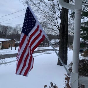 May include: A United States flag featuring a blue canton with white stars and alternating red and white horizontal stripes, mounted on a white pole attached to a white porch column, displayed outdoors during a winter day with snow on the ground.