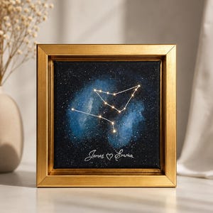 Può includere: Un quadro su tela quadrato in una cornice dorata che raffigura un cielo notturno blu scuro e nero con una costellazione luminosa. I nomi James ed Emma sono scritti in corsivo bianco in basso, separati da un piccolo simbolo a cuore.