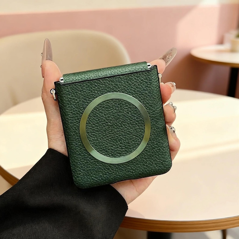 K&ouml;nnte beinhalten: Eine dunkelgr&uuml;ne Handyh&uuml;lle mit genarbter Lederoptik und einem markanten, mittig platzierten Metallring. Das rechteckige Etui hat abgerundete Ecken und verf&uuml;gt &uuml;ber kleine Metall&ouml;sen an den oberen Seiten zur Befestigung eines Trageriemens.