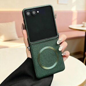 K&ouml;nnte beinhalten: Eine dunkelgr&uuml;ne, texturierte Handyh&uuml;lle in Lederoptik f&uuml;r ein faltbares Smartphone. Die H&uuml;lle verf&uuml;gt &uuml;ber einen kreisf&ouml;rmigen Magnetring auf der R&uuml;ckseite und kleine Metall&ouml;sen an der Seite zur Befestigung eines Riemens. Zwei Kameralinsen.