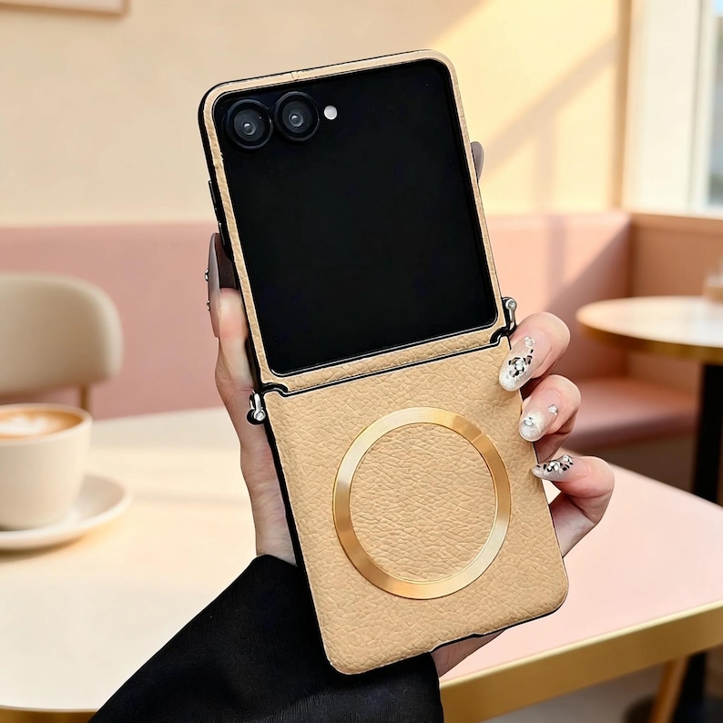 K&ouml;nnte beinhalten: Eine beige, texturierte Handyh&uuml;lle f&uuml;r ein faltbares Smartphone mit einem markanten goldenen, kreisf&ouml;rmigen Magnetring auf der unteren R&uuml;ckseite. Die H&uuml;lle hat einen schwarzen Rahmen und Aussparungen f&uuml;r die Doppelkamera und den Blitz.