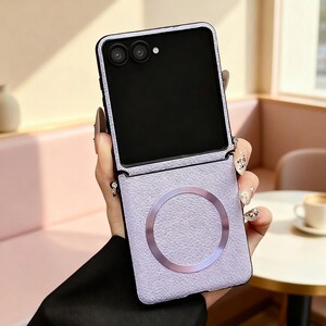 K&ouml;nnte beinhalten: Eine faltbare Smartphone-H&uuml;lle in hellem Lavendel mit einer strukturierten, genarbten Lederoptik. Die H&uuml;lle verf&uuml;gt &uuml;ber einen markanten, metallischen kreisf&ouml;rmigen Ring in der Mitte der unteren H&auml;lfte, der f&uuml;r magnetische Befestigungen gedacht ist.
