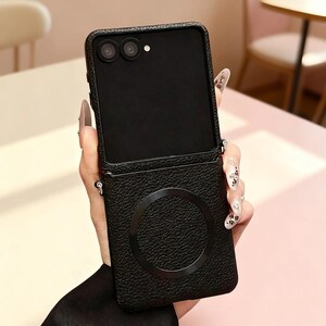 K&ouml;nnte beinhalten: Eine schwarze, faltbare Smartphone-H&uuml;lle mit einer strukturierten, lederartigen Oberfl&auml;che. Die H&uuml;lle verf&uuml;gt &uuml;ber einen kreisf&ouml;rmigen magnetischen Ladering auf der unteren R&uuml;ckseite und zwei kreisf&ouml;rmige Kameraaussparungen oben.
