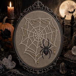 May include: An oval-framed embroidery piece featuring a white beaded spiderweb on a tan fabric background. A black beaded spider with a red accent sits on the web. Gold-toned beaded chains hang from the web, all encased in an ornate dark frame.
