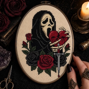 Könnte beinhalten: Ein ovaler Stickrahmen mit einem Kreuzstichmotiv einer Kapuzenfigur mit weißer Maske, die ein rotes Herz hält, das von einem silbernen Schwert durchbohrt wird, umgeben von roten und schwarzen Rosen auf einem cremefarbenen Stoffhintergrund.