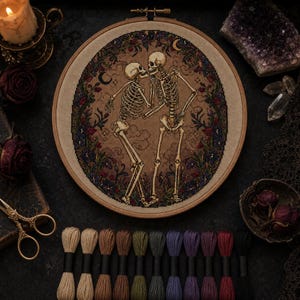 May include: A circular wooden embroidery hoop containing a cross-stitch design of two skeletons dancing. The skeletons are surrounded by a dark floral wreath with crescent moons and stars on a tan fabric background, accompanied by embroidery floss.