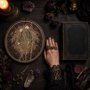 May include: A circular wooden embroidery hoop containing a cross-stitch design of two skeletons facing each other, surrounded by a floral wreath with crescent moons. The embroidery is set against a dark background with a black book and scissors.