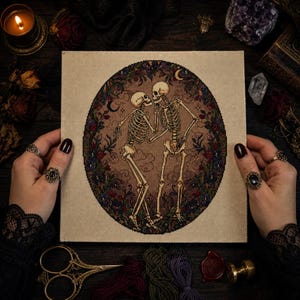 May include: A square fabric piece featuring a cross-stitch embroidery of two skeletons dancing within a circular floral border. The design includes dark red and blue flowers, crescent moons, and a brown background, all stitched on light beige fabric.
