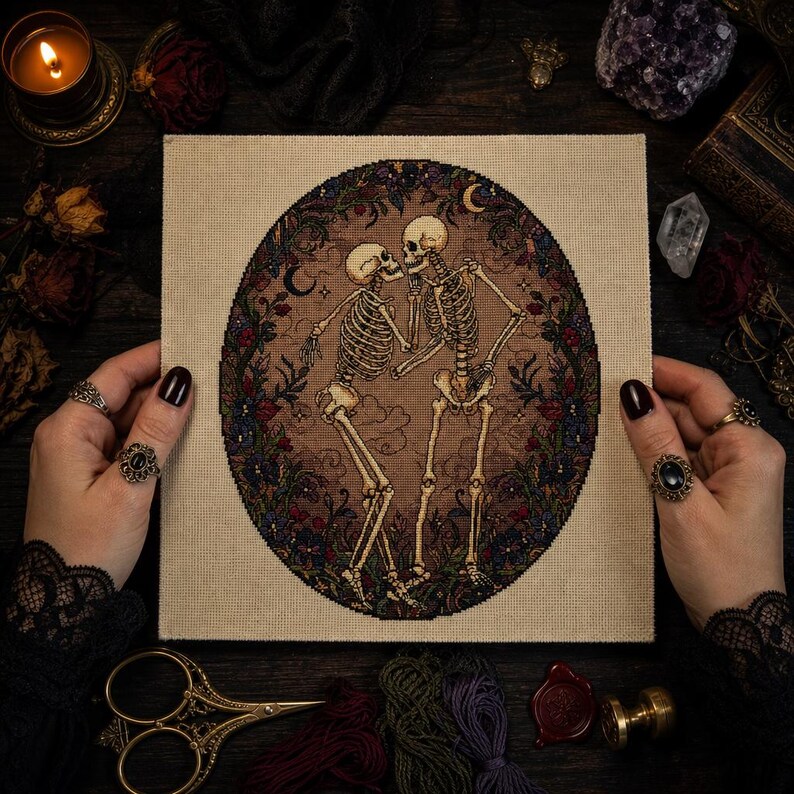 May include: A square fabric piece featuring a cross-stitch embroidery of two skeletons dancing within a circular floral border. The design includes dark red and blue flowers, crescent moons, and a brown background, all stitched on light beige fabric.