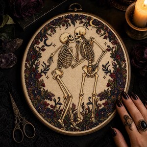 Op de afbeelding: Een cirkelvormig kruissteek borduurwerk met twee dansende skeletten, ingelijst in een houten ring. De skeletten zijn omringd door een gedetailleerde rand van donkerpaarse, rode en groene bloemen, bladeren en kleine maansikkels op beige stof.